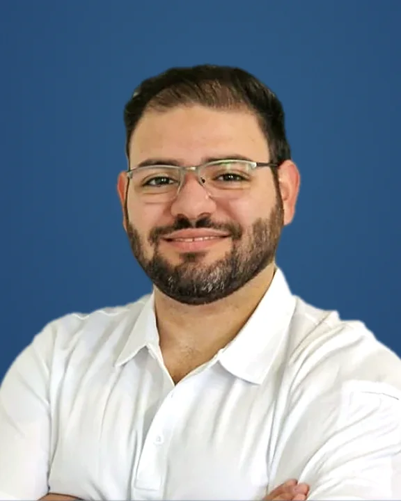Porträt von Dr. Bashar Kahwaji mit Brille und Bart im weißen Poloshirt mit verschränkten Armen vor blauem Hintergrund, lächelnd.
