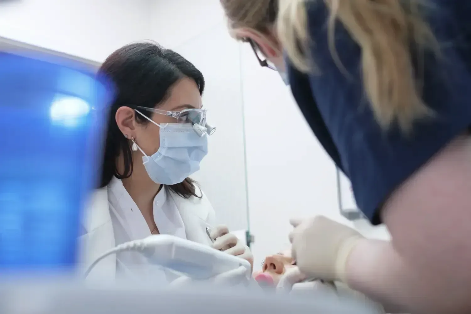 Eine Zahnärztin mit Mundschutz und Lupenbrille behandelt eine Patientin. Von rechts beugt sich eine Mitarbeiterin ins Bild.