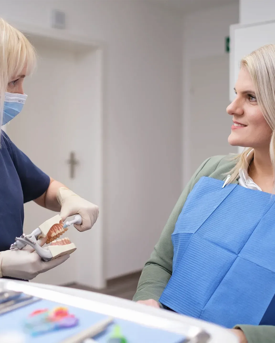 Eine Mitarbeiterin erklärt einer Patientin im Behandlungsstuhl die Zahnpflege an einem Kiefermodell. Die Patientin trägt ein blaues Lätzchen und hört aufmerksam zu.