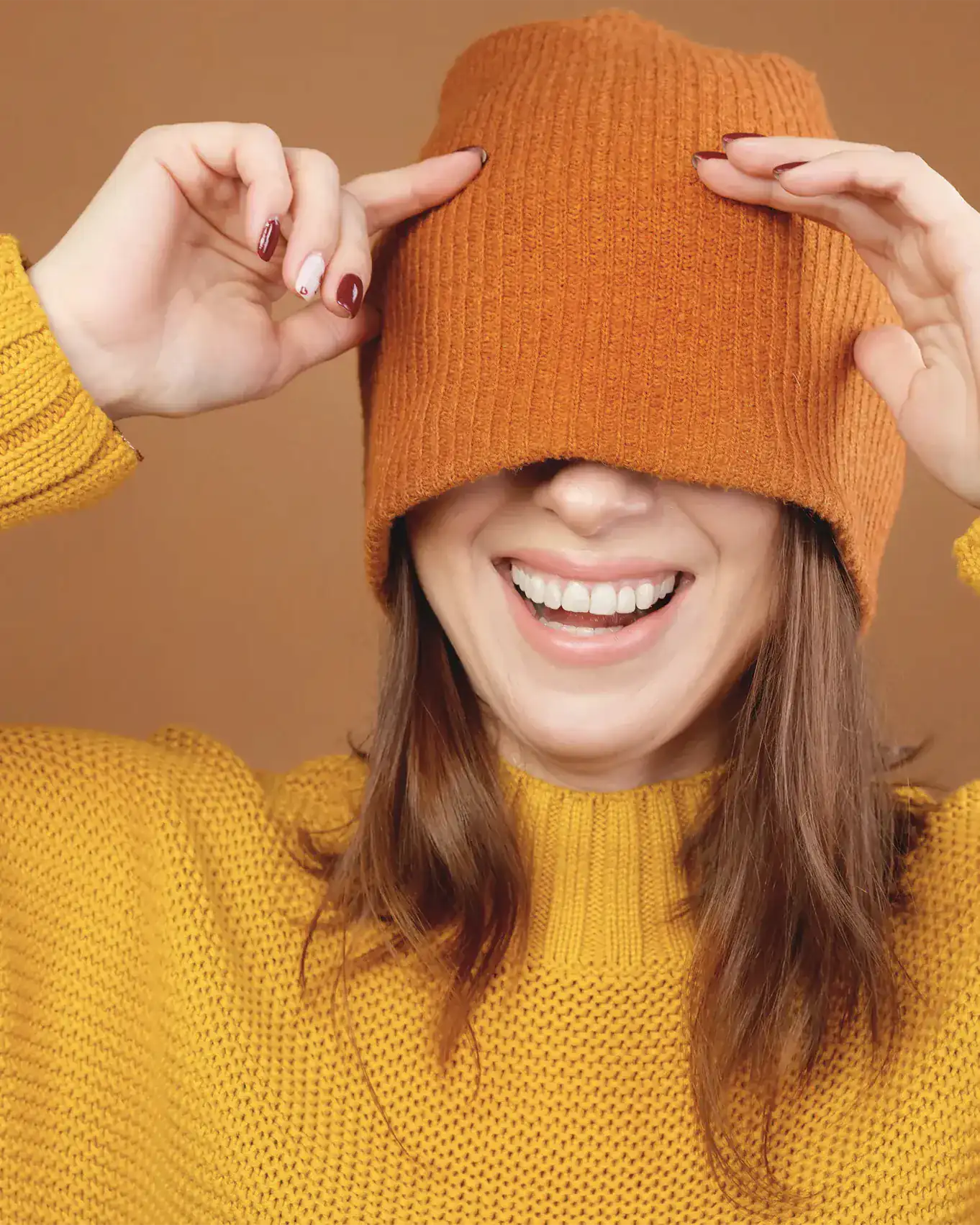 Eine Frau im gelben Pullover zieht sich eine orange Mütze über die Augen und lacht, ihre Zähne sind zu sehen. Symbolbild.