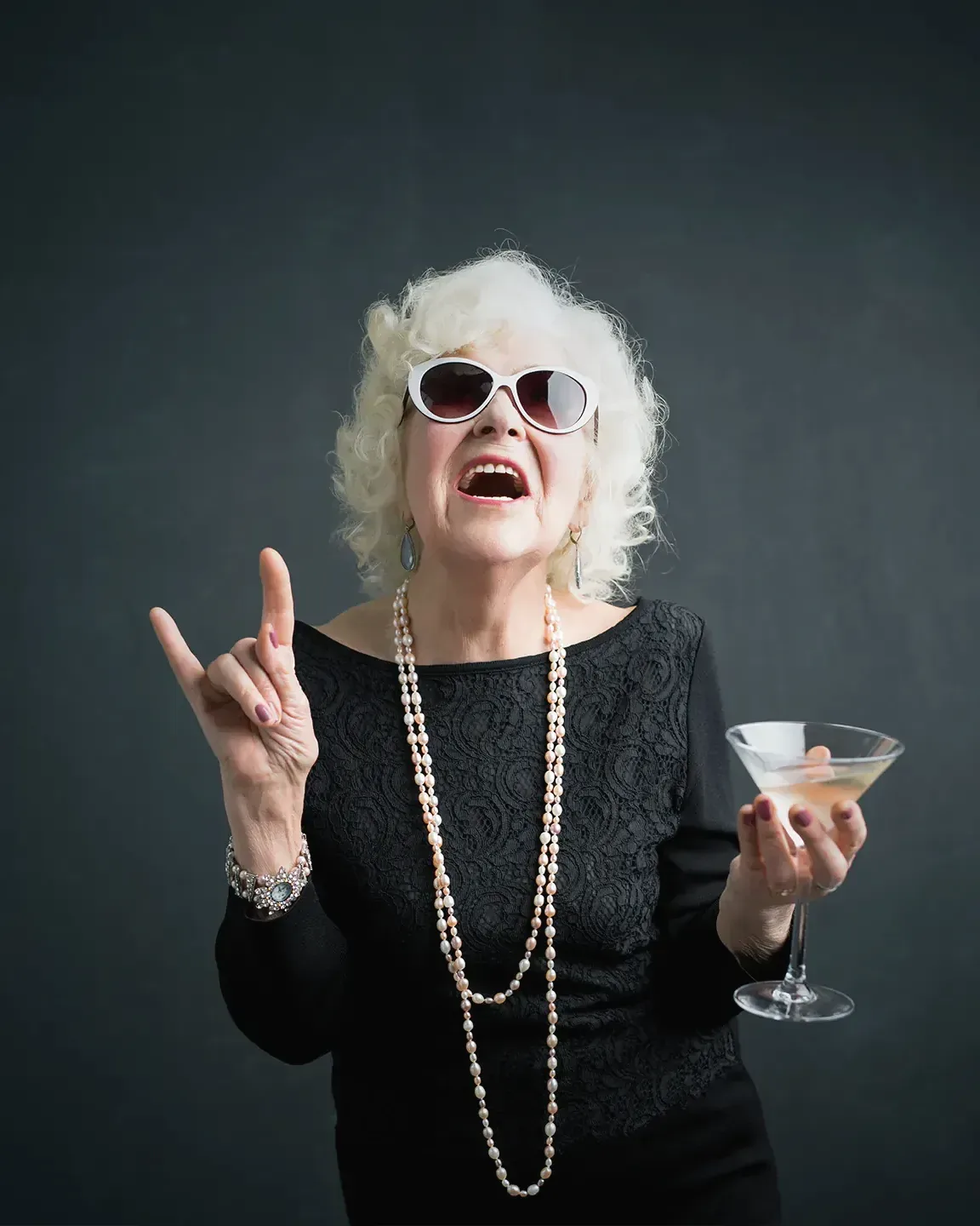 Eine ältere Frau mit weißen Locken, Sonnenbrille und Perlenkette lacht ausgelassen und hält ein Glas. Symbolbild.