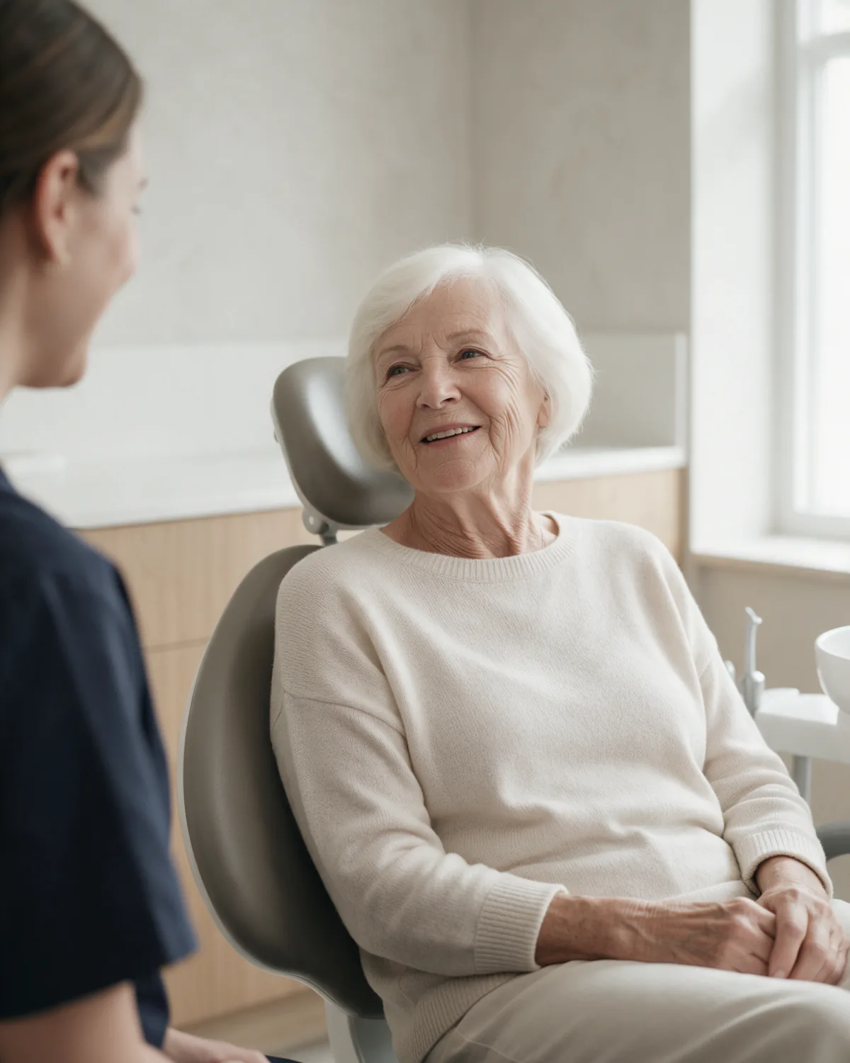 Eine Frau um die fünfundsiebzig mit kurzen weißen Haaren sitzt entspannt im Behandlungsstuhl und unterhält sich freundlich mit einer Mitarbeiterin, die am linken Bildrand angeschnitten ist. KI-generiertes Symbolbild.