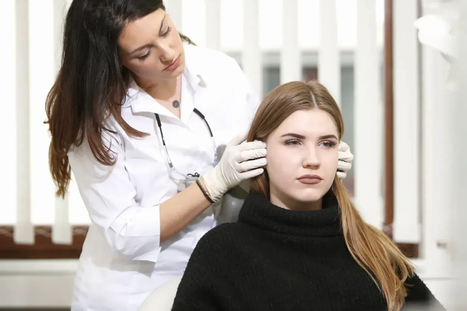 Eine Zahnärztin im weißen Kittel tastet mit Handschuhen vor den Ohren einer jungen Patientin die Kiefergelenke ab. Die Patientin schaut ernst geradeaus.