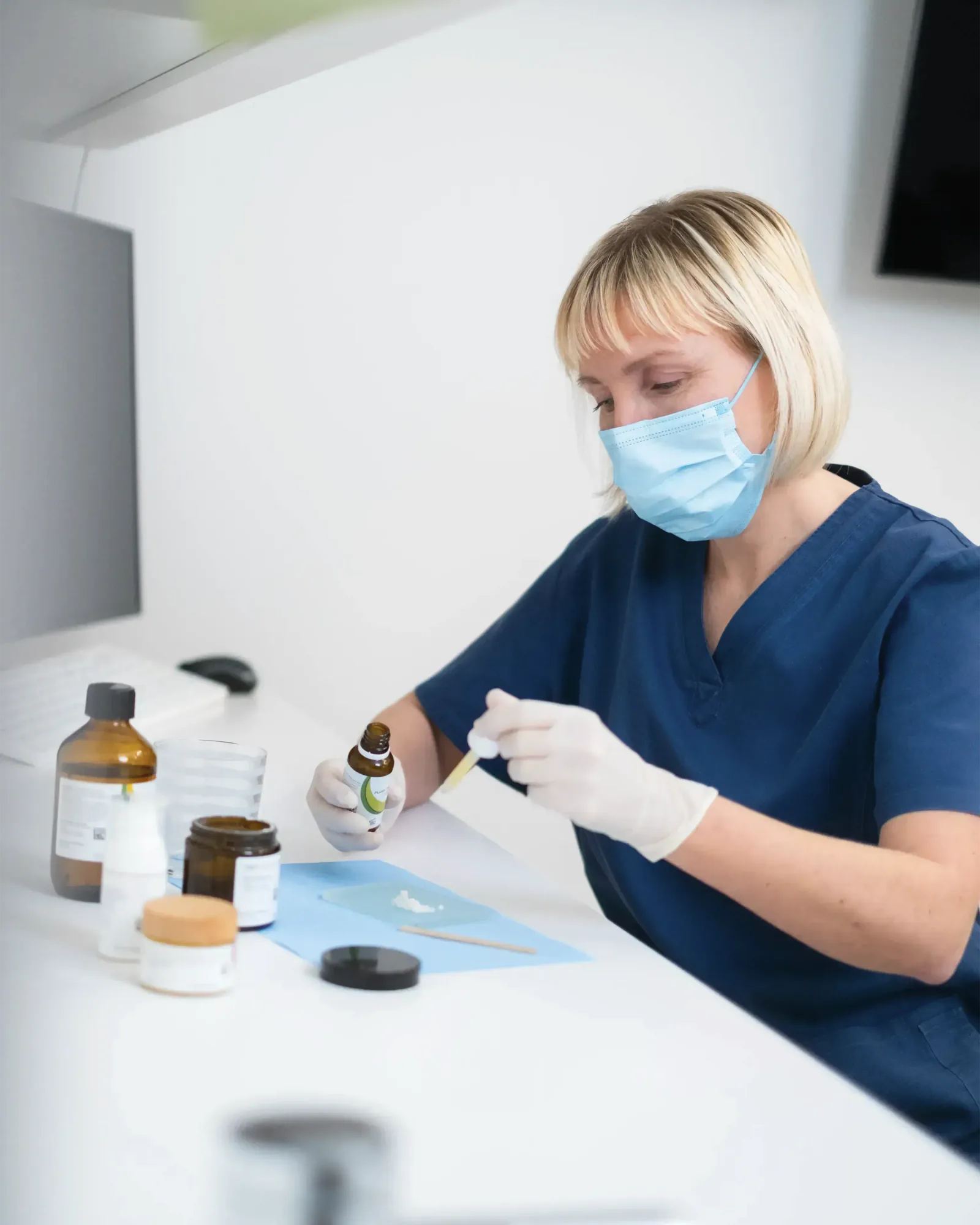 Eine Mitarbeiterin mit Mundschutz tropft am Schreibtisch aus einem kleinen Fläschchen mit einer Pipette Flüssigkeit auf eine Mischplatte. Links stehen Braunglasflaschen und Döschen.