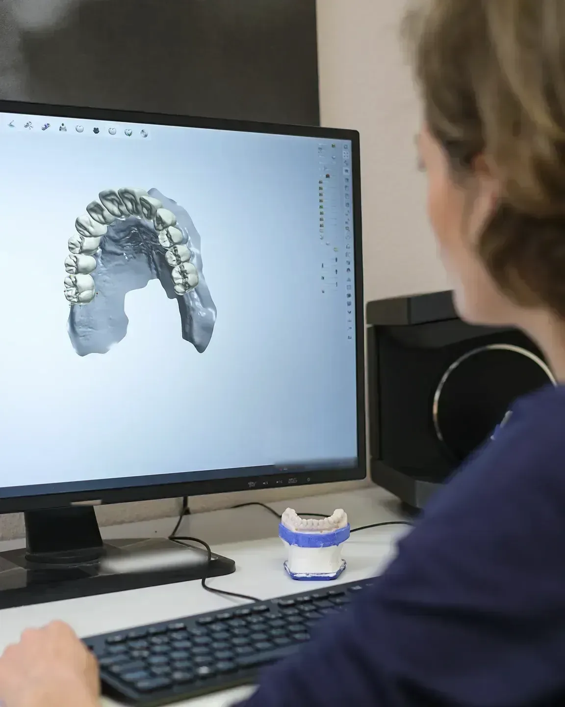 Eine Mitarbeiterin sitzt von der Seite am Schreibtisch vor einem Bildschirm, der das digitale Modell eines Kiefers zeigt. Auf dem Tisch steht ein Gipsmodell.