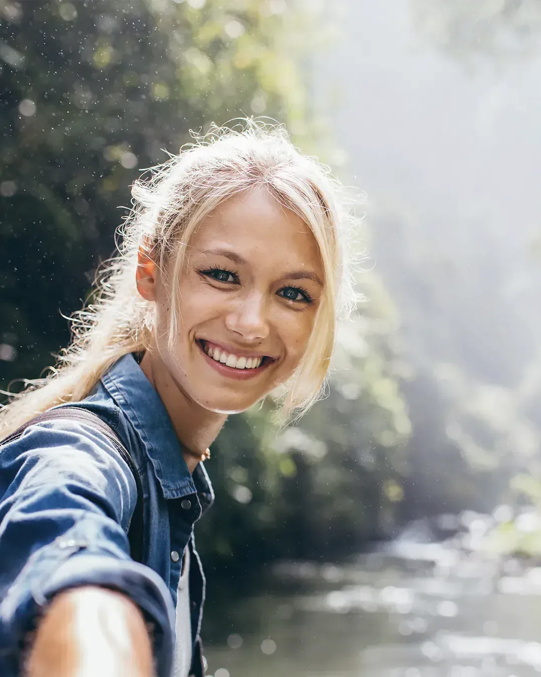 Eine junge Frau mit blonden Haaren lächelt an einem Fluss im Wald in die Kamera, Morgenlicht fällt durch die Bäume. Symbolbild.