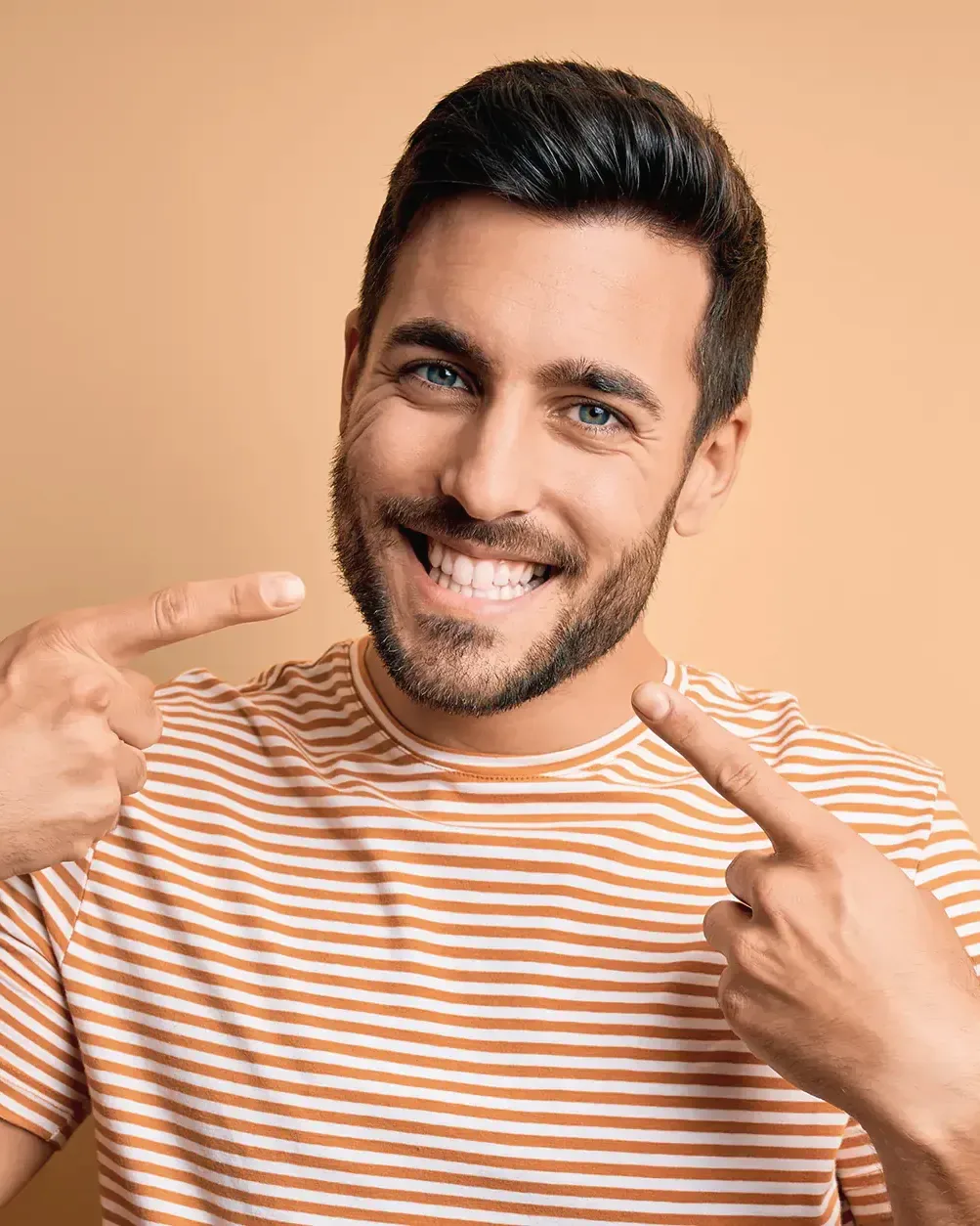 Ein junger Mann mit Bart und gestreiftem T-Shirt lächelt breit und zeigt mit beiden Zeigefingern auf sein Lächeln, vor orangefarbenem Hintergrund. Symbolbild.
