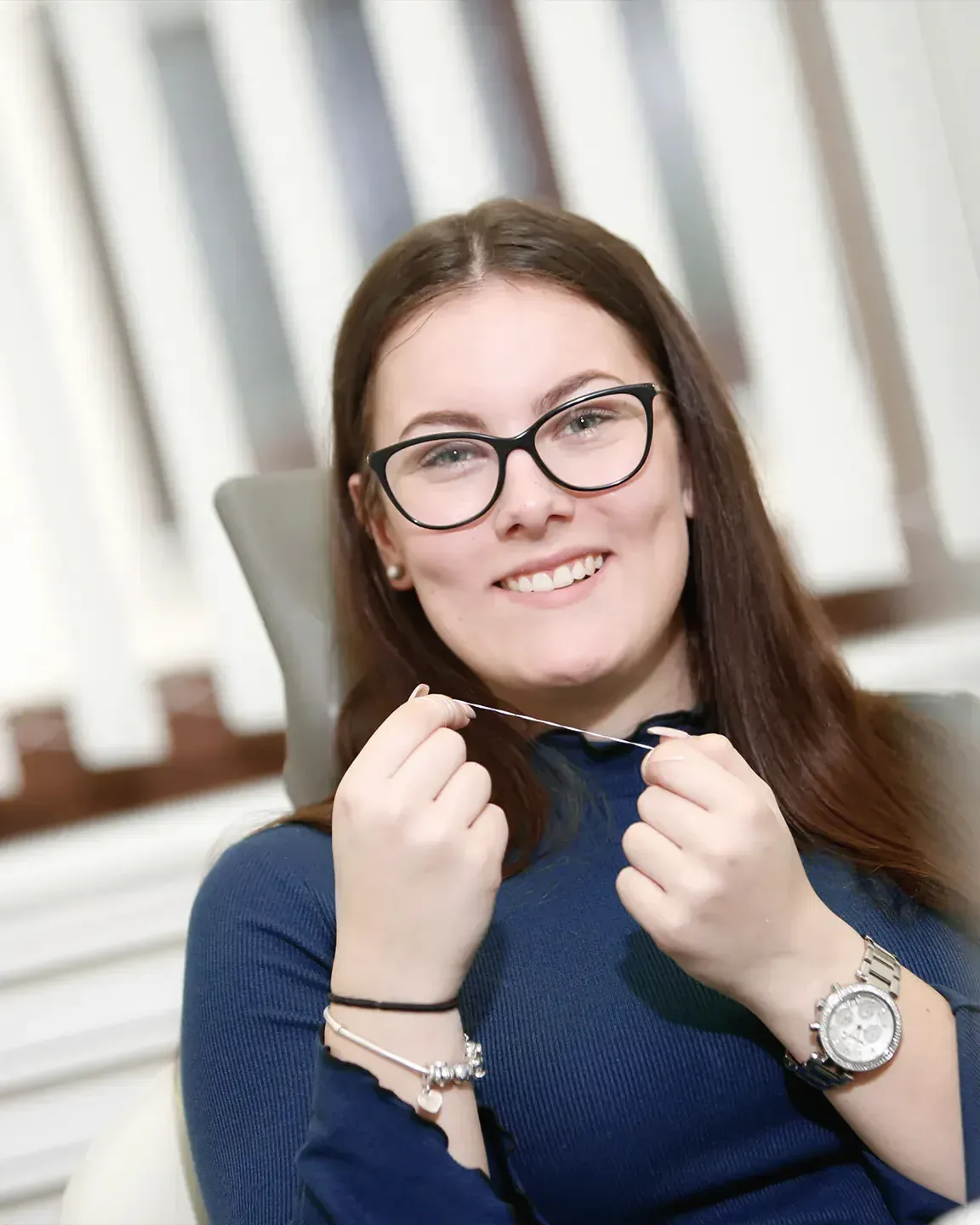 Eine junge Patientin mit Brille sitzt im Behandlungsstuhl, hält Zahnseide zwischen den Händen und lächelt.